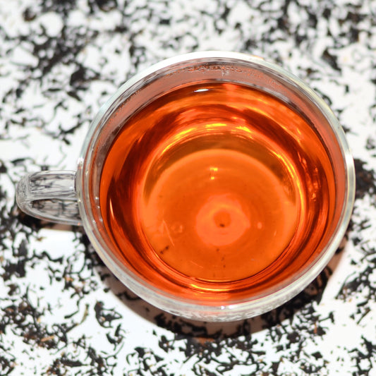 Photograph of Easy Assam TGFOP (Tippy Golden Flowery Orange Pekoe) Tea Brewed in a glass cup with loose leaves scattered in the background. The brewed tea is golden-brown and vibrant in colour.