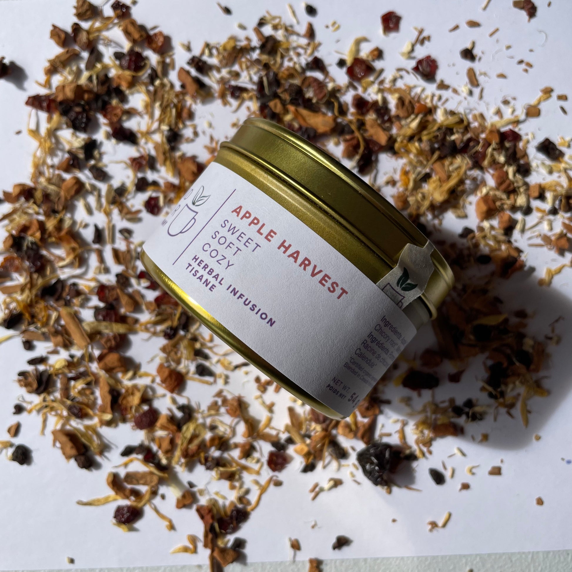Island Elixir Apple Harvest herbal tea 'Essential Tin' size package surrounded by loose tea leaves on a white background