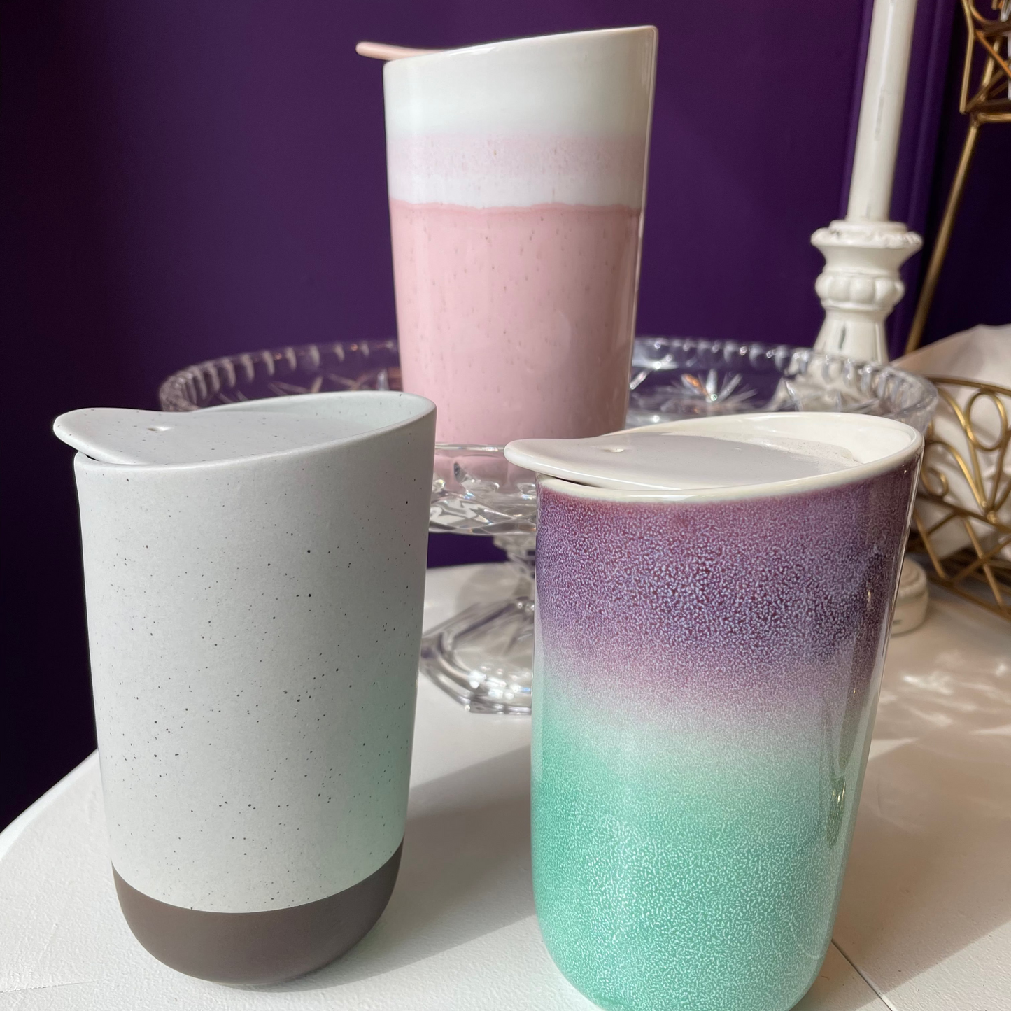 Three ceramic tumblers with integrated lids are displayed on a white surface against a dark purple background. The tumblers are available in theee colours: a light brown with textural speckle glaze in a matte finish; a white and dusty pink with white lid on a smooth glossy finish; a purple and turquoise with a white lid and a smooth glossy finish. Perfect for tea on the go (also good for coffee or hot chocolate). 