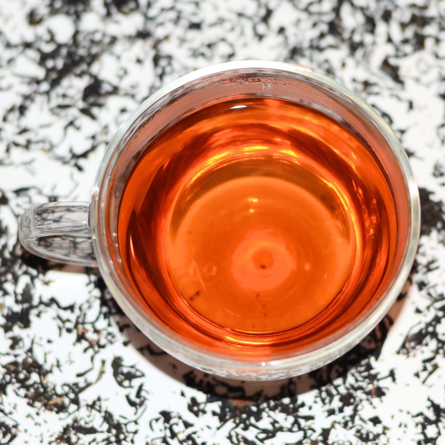 Photograph of Easy Assam TGFOP (Tippy Golden Flowery Orange Pekoe) Tea Brewed in a glass cup with loose leaves scattered in the background. The brewed tea is golden-brown and vibrant in colour.
