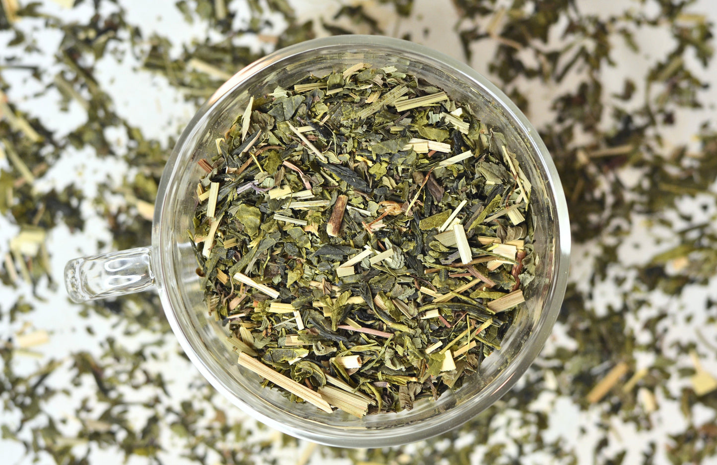 Glass teacup filled with Ingenuitea green tea blend loose tea leaves. Tea leaves scattered in the background.