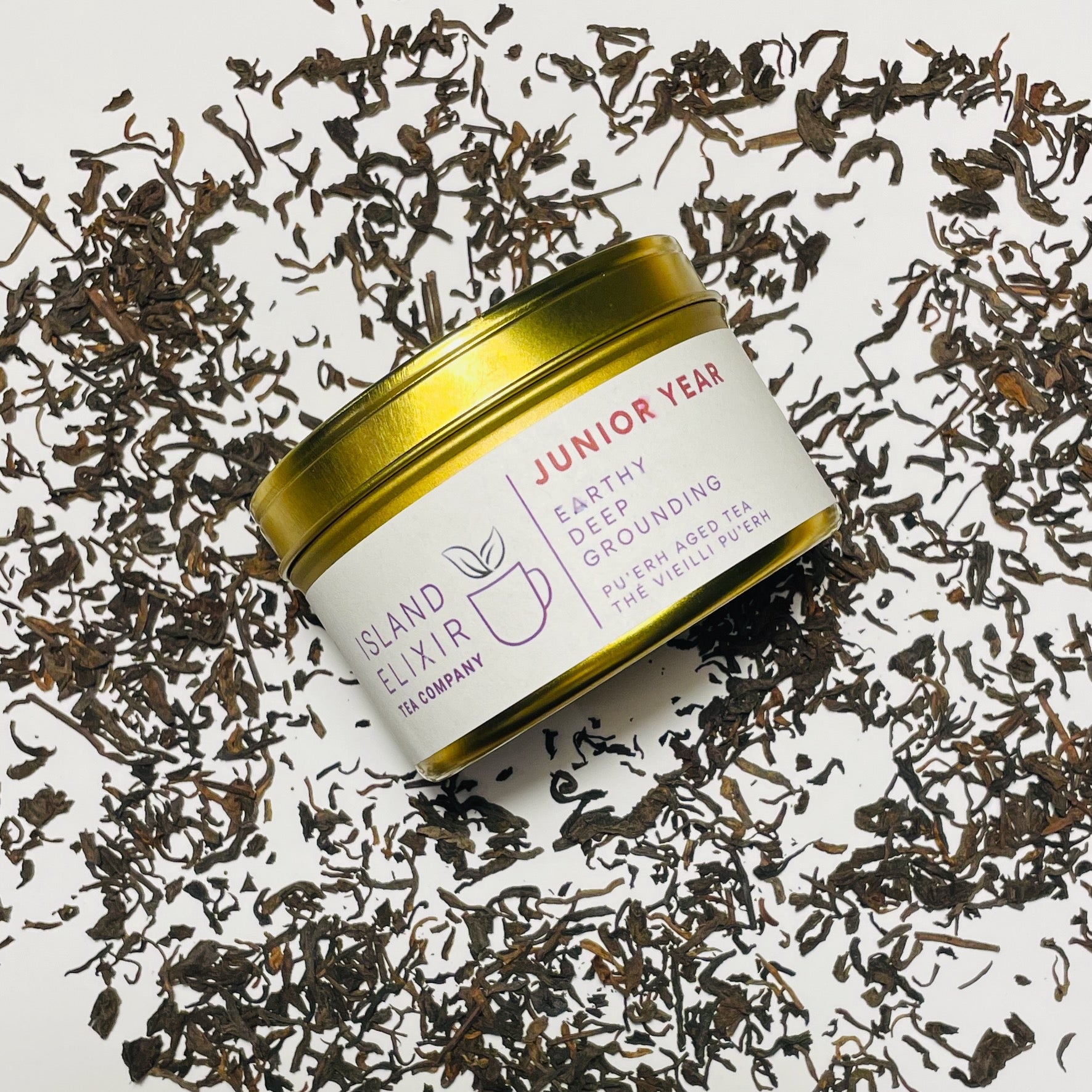 Junior Year Puerh dark tea in gold tin sitting on top of scattered tea leaves on a white background