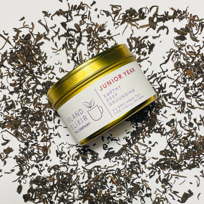 Junior Year Puerh dark tea in gold tin sitting on top of scattered tea leaves on a white background