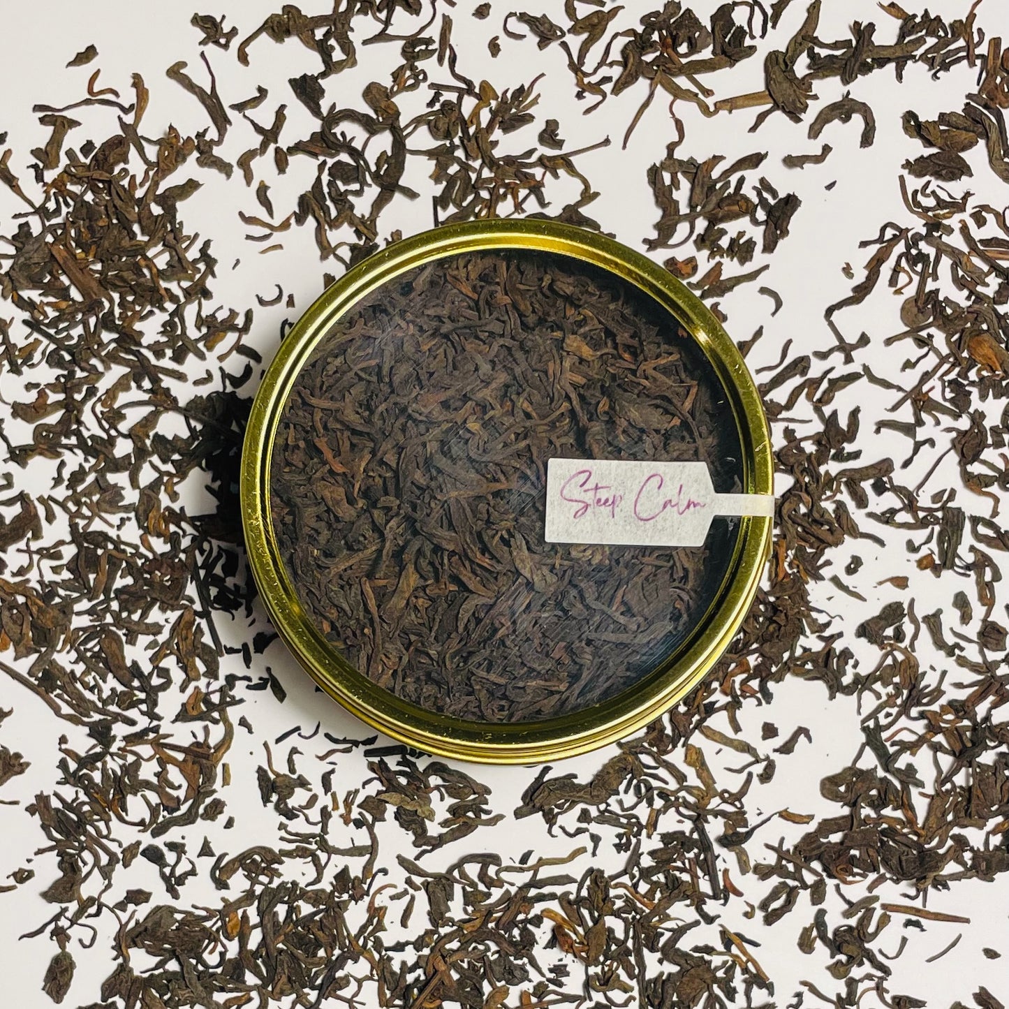 Junior Year Puerh dark tea in gold tin viewed from the top sitting on top of scattered tea leaves on a white background