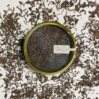 Junior Year Puerh dark tea in gold tin viewed from the top sitting on top of scattered tea leaves on a white background