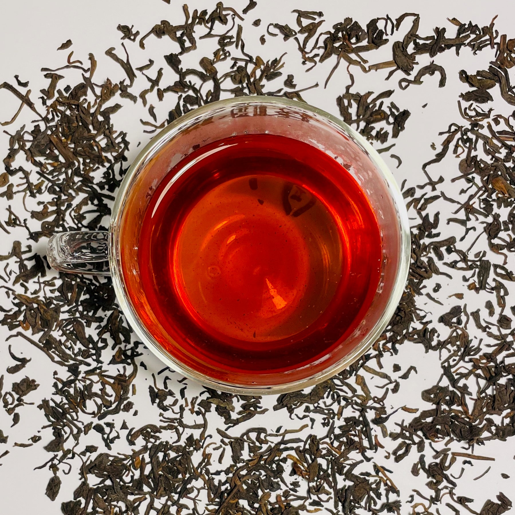 Junior Year Puerh dark tea in a glass cup on  top of scattered tea leaves on a white background