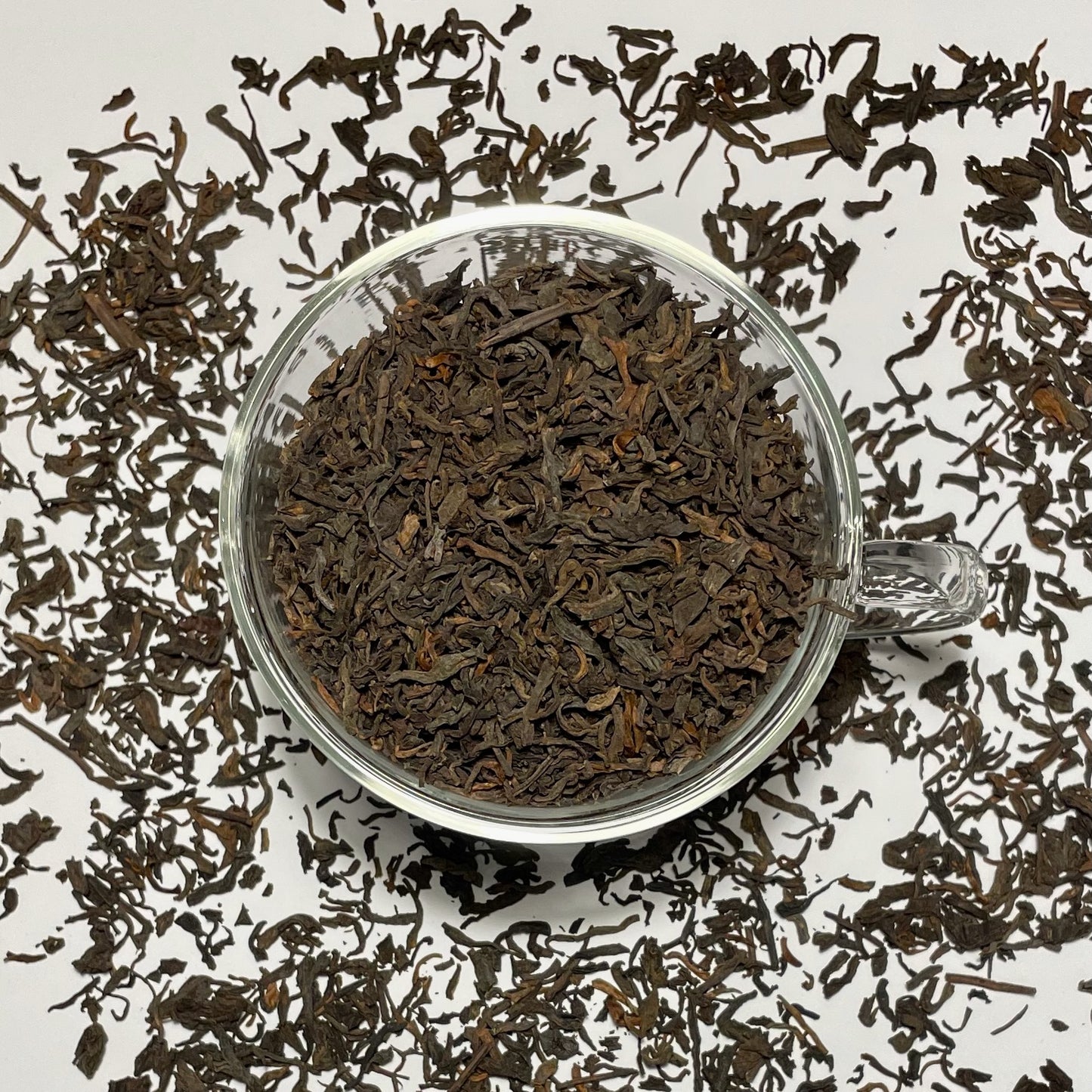 Junior Year Puerh dark tea leaves in a glass cup sitting on top of scattered tea leaves on a white background