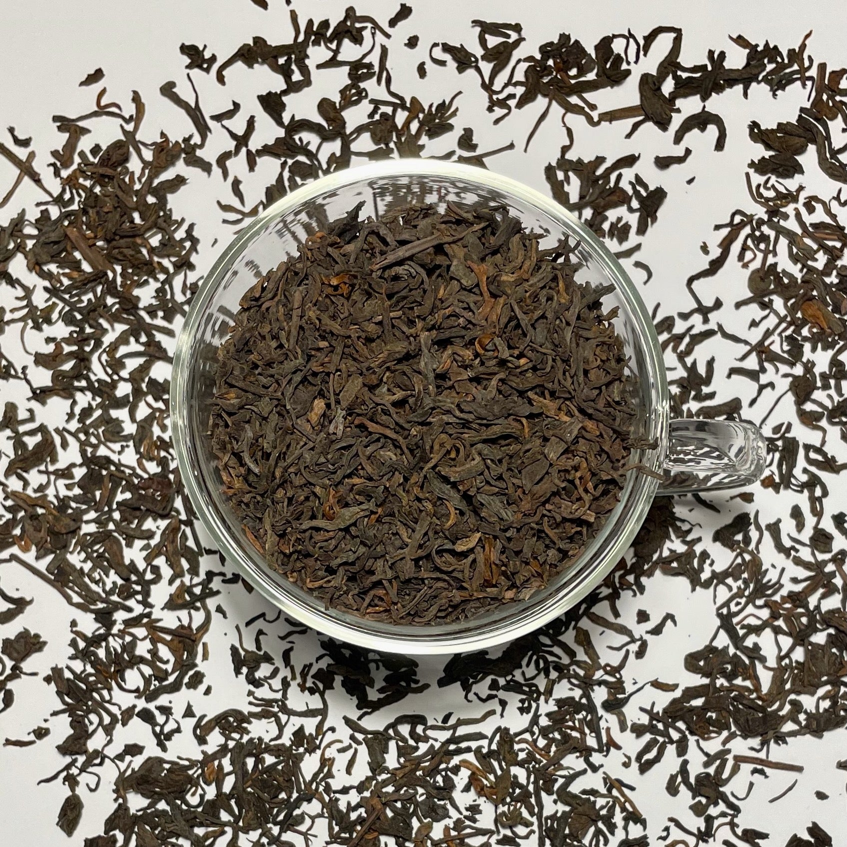 Junior Year Puerh dark tea leaves in a glass cup sitting on top of scattered tea leaves on a white background