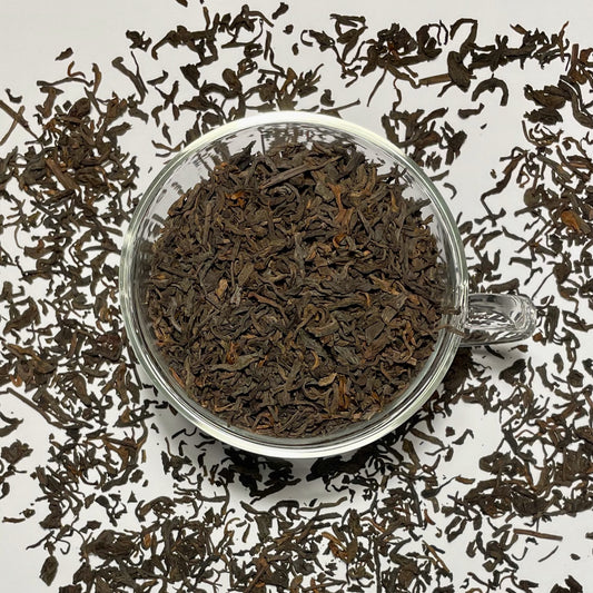 Junior Year Puerh dark tea leaves in a glass cup sitting on top of scattered tea leaves on a white background