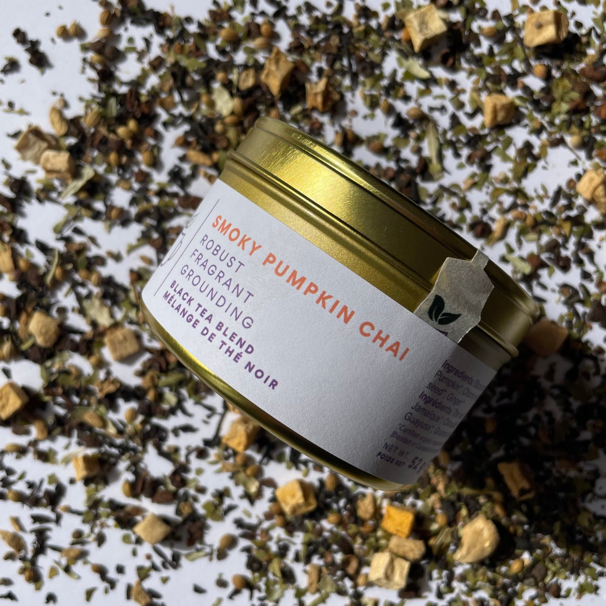 Island Elixir Smoky Pumpkin Chai tea 'Essential tin' size package viewed from the side and surrounded by loose tea leaves on a white background