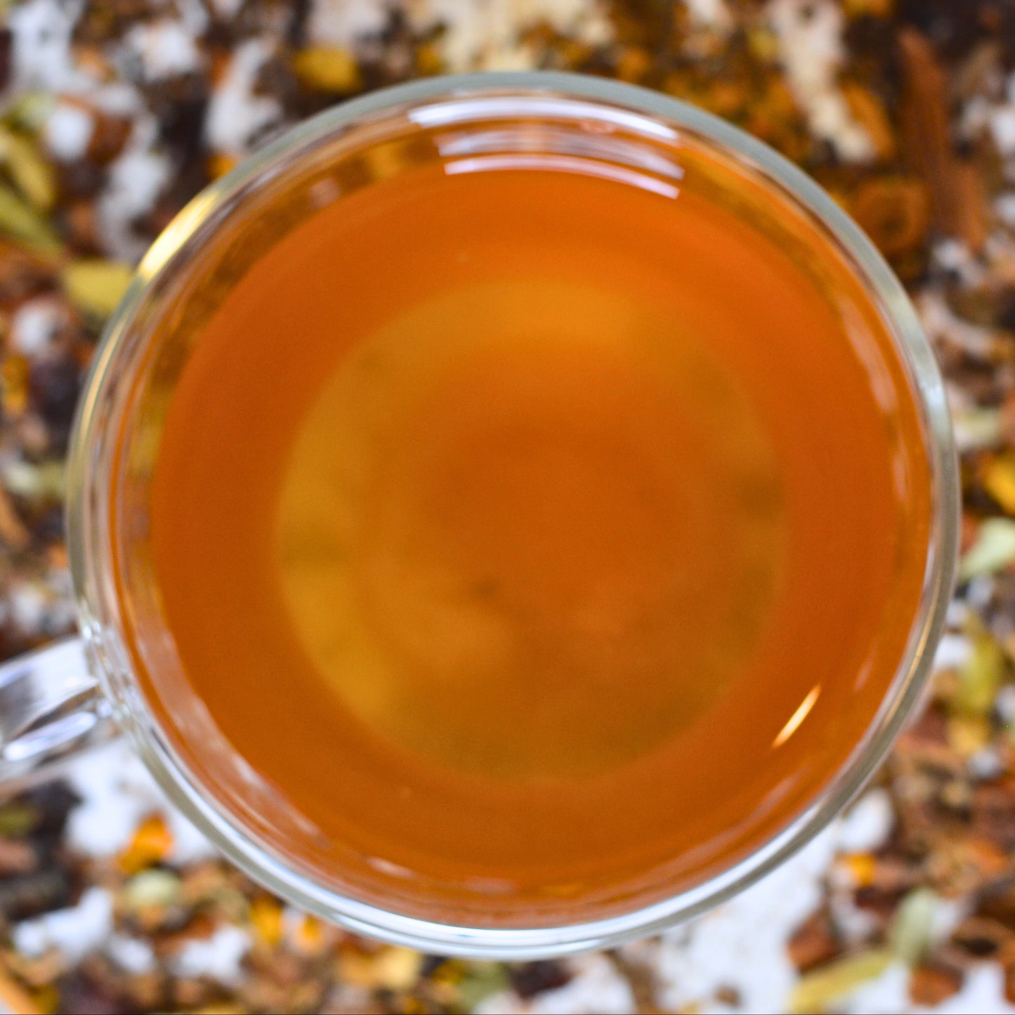 Sumitra's Chai Tea Brewed. Steeped chai in a glass cup showing the golden orange colour. Loose leaf tea blurred in the background.