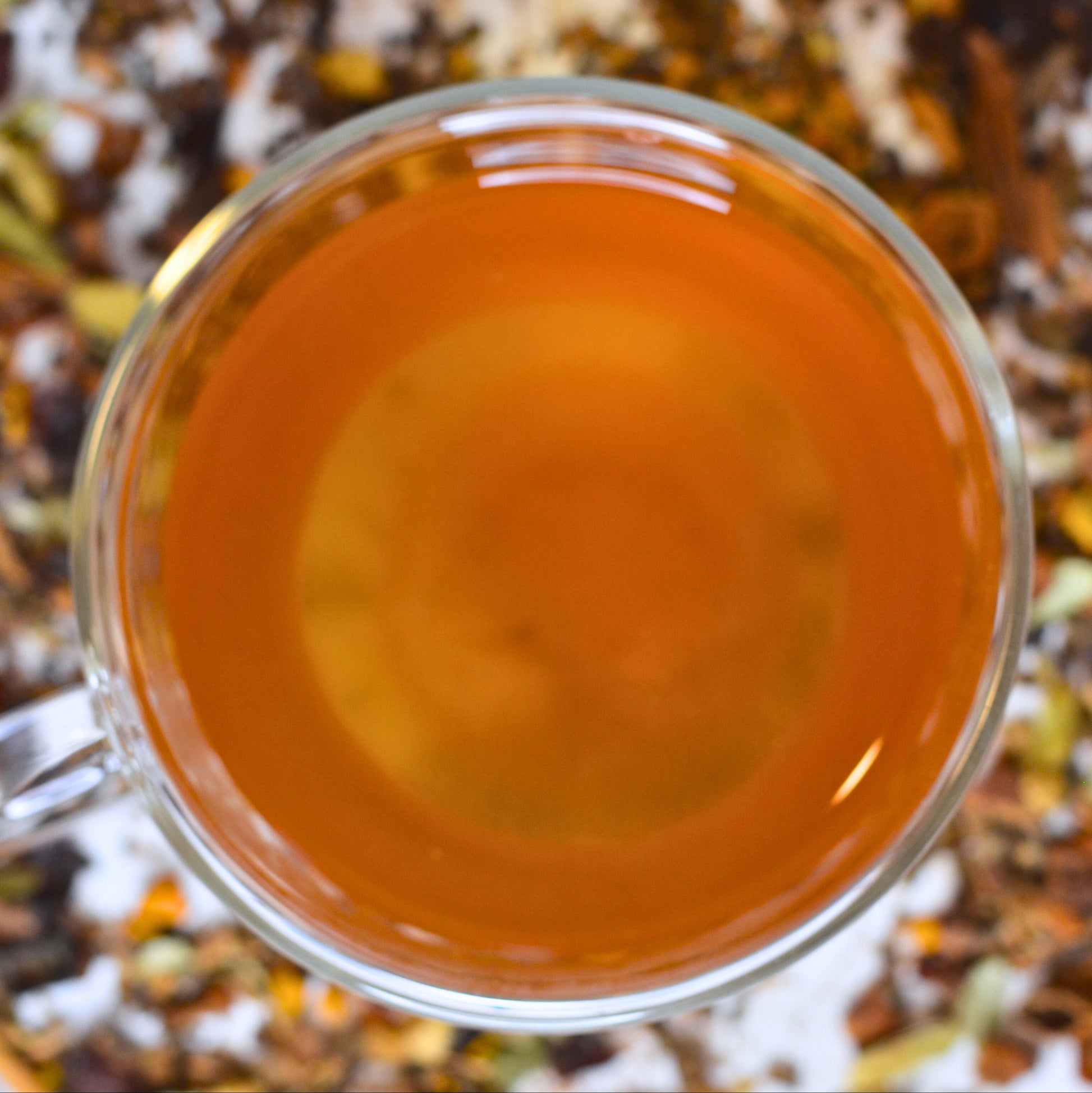 Sumitra's Chai Tea Brewed. Steeped chai in a glass cup showing the golden orange colour. Loose leaf tea blurred in the background.