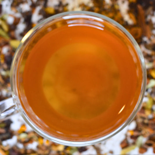 Sumitra's Chai Tea Brewed. Steeped chai in a glass cup showing the golden orange colour. Loose leaf tea blurred in the background.