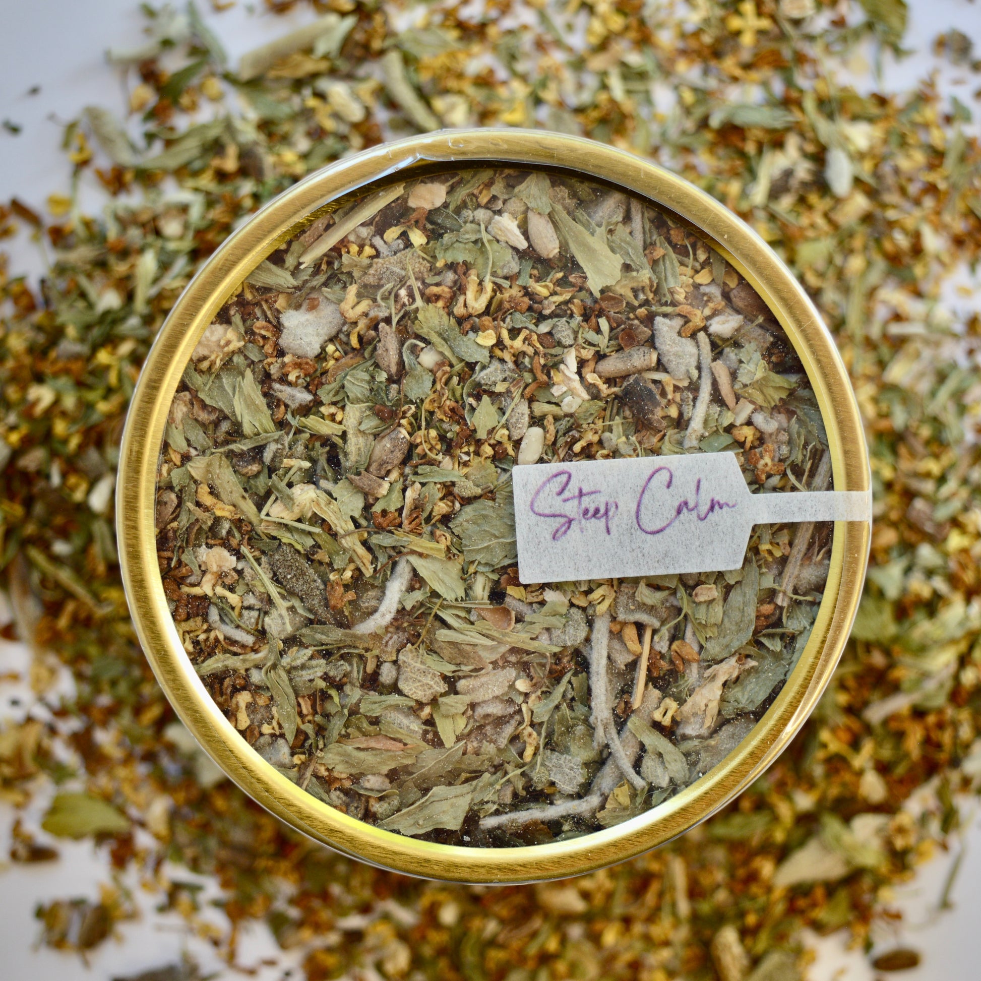 The Legends Herbal Tea 30 cups of tea size gold finish tin sitting on loose tea that contains Milk thistle seed, Osmanthus, Dandelion root, Alfalfa, Sage, Rosemary.