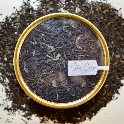 Top view Black Dragon Black Tea 30 cups of tea size gold finish tin sitting on loose leaf tea that contains Black tea*, Jasmine scented green tea*.