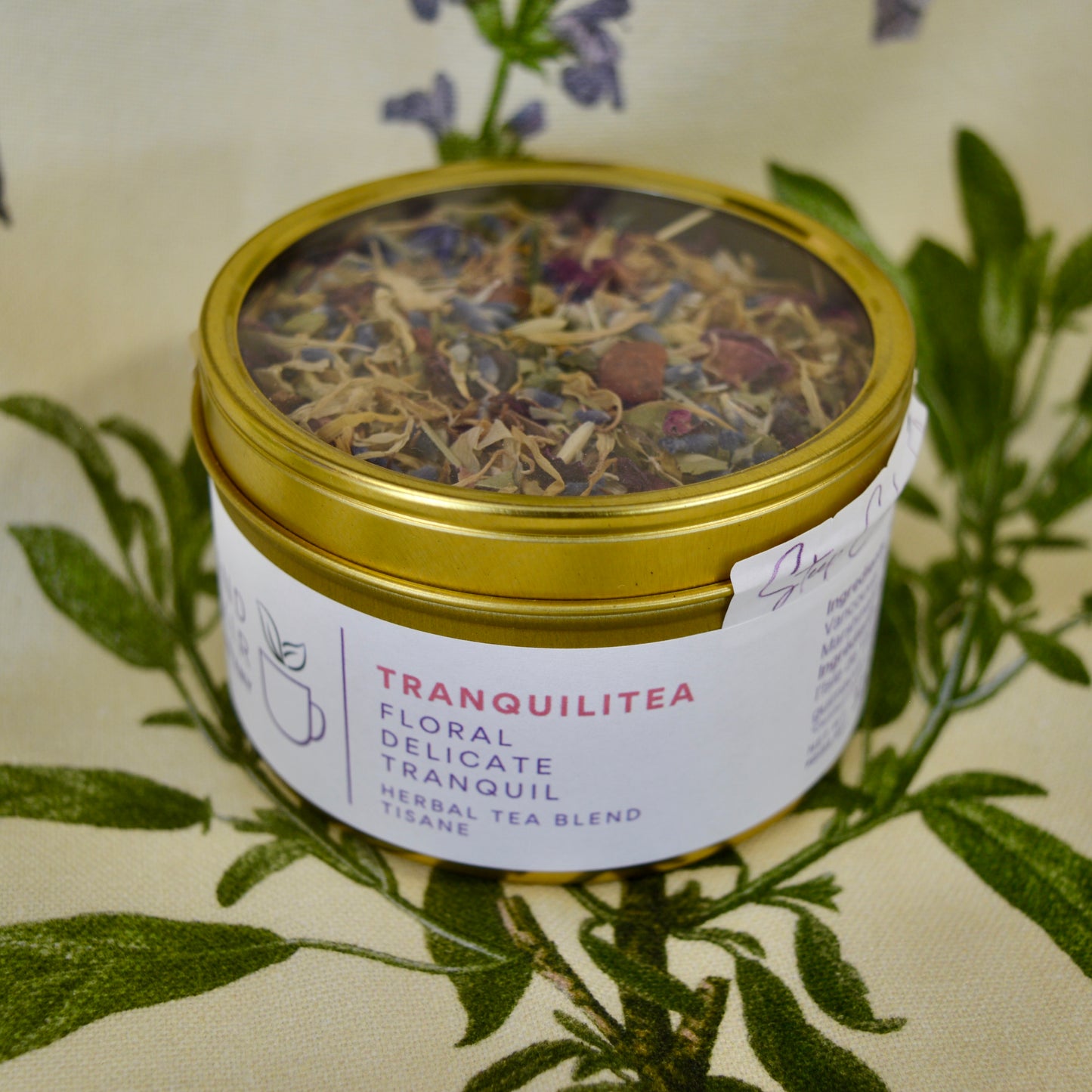 Tranquilitea Herbal Tea 30 cups of tea size gold finish tinsitting on loose leaf tea that contains Rose hip*, Lemon balm*, Lavender**, Oatstraw*, Marshmallow leaf*, Calendula*, Rose petals*.